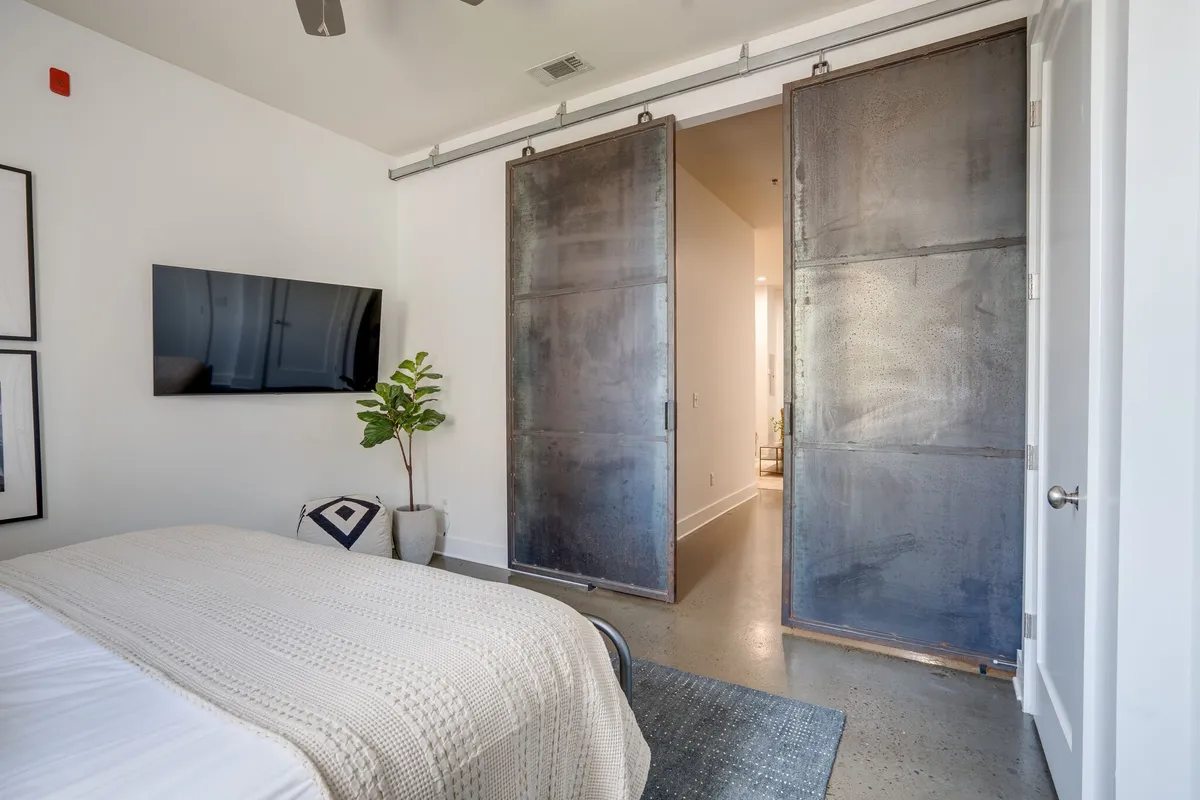 A tranquil bedroom highlighted by sliding barn doors, a bed with a textured blanket, a television opposite the bed, and a potted plant in the corner. Soft natural light enters the room, enhancing the sense of peaceful retreat and flexibility.