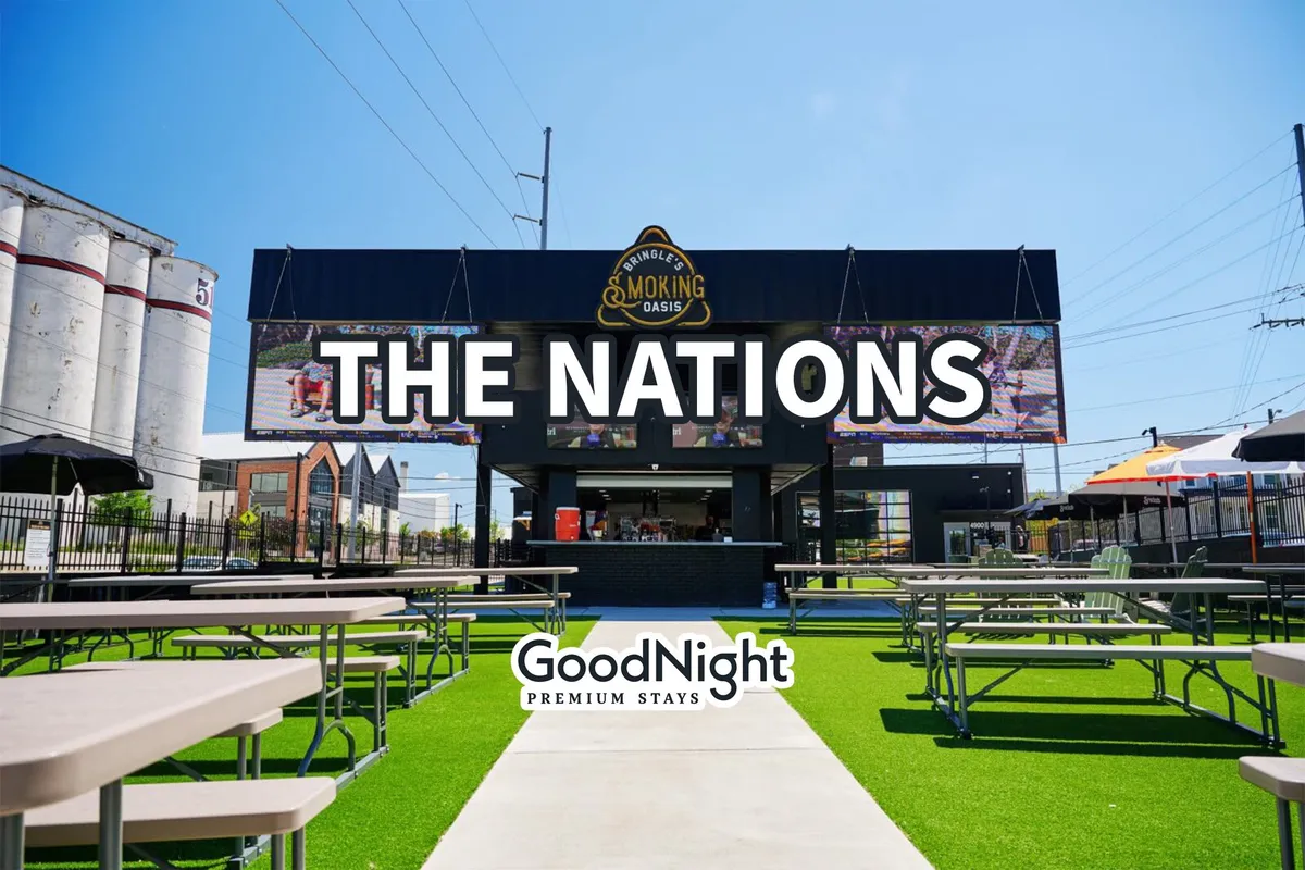 A vibrant exterior scene featuring a large sign reading 'THE NATIONS', outdoor seating, green grass, and people enjoying food and community events. The atmosphere is energetic and welcoming, ideal for social gatherings and outdoor family activities.