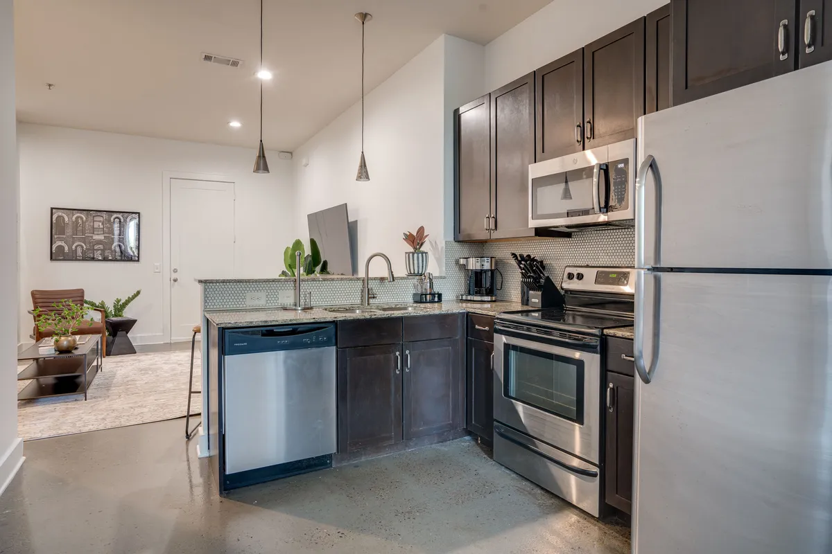 A modern kitchen featuring sleek stainless steel appliances, dark cabinetry, and a textured countertop. Bar seating lines the countertop, and a decorative plant adds a touch of vibrancy. The kitchen is open and inviting, ideal for cooking and entertaining.