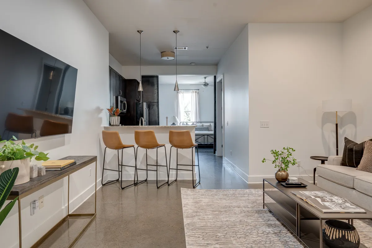 A contemporary open-plan interior displaying a modern sofa, bar stools at a kitchen island, a TV unit, and a central coffee table. The flow between kitchen and living space is obvious, perfectly supporting group mingling and shared moments.