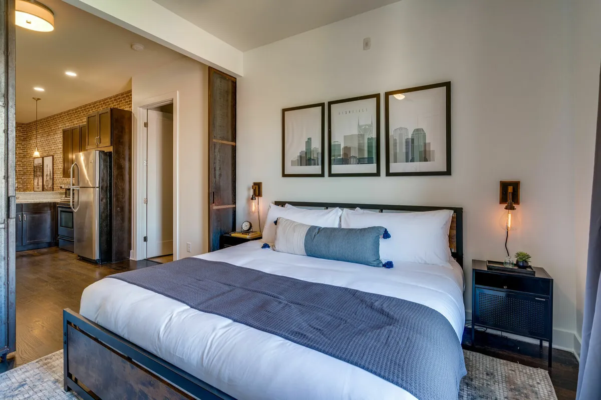A cozy king bedroom featuring a large bed with plush bedding, soft throw pillows, a framed cityscape artwork above the headboard, and a nightstand with a contemporary lamp. Natural light filters into the room, creating a serene and inviting atmosphere.