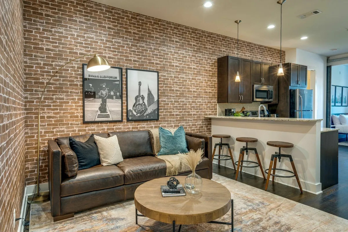 The image showcases a stylish, open-concept living area with an exposed brick wall, contemporary artwork, a plush sofa perfect for lounging, and a modern kitchen island inviting guests to gather. The space feels cozy and artistic, ideal for relaxing or entertaining with friends.