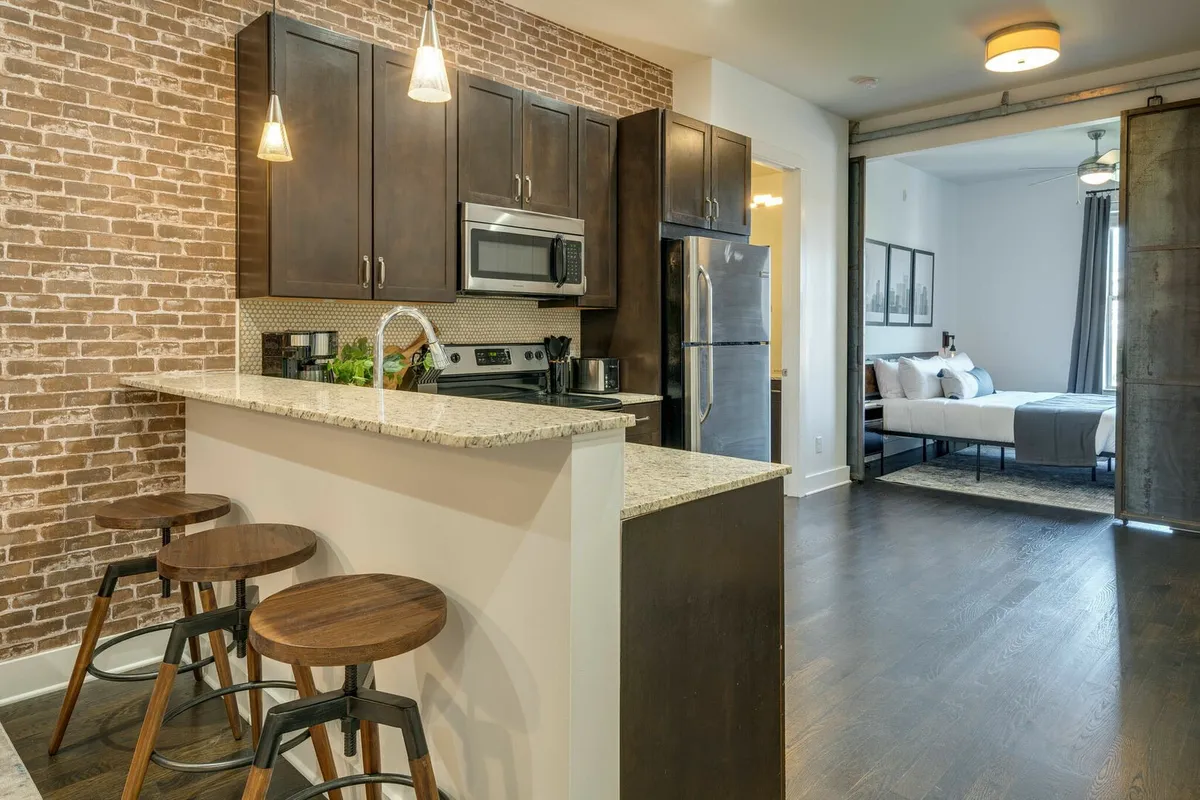 A cozy kitchen area with a central island, exposed brick wall, bar stools, and modern appliances. The kitchen opens to a living area, enhancing the sense of communal space for cooking, eating, and socializing.