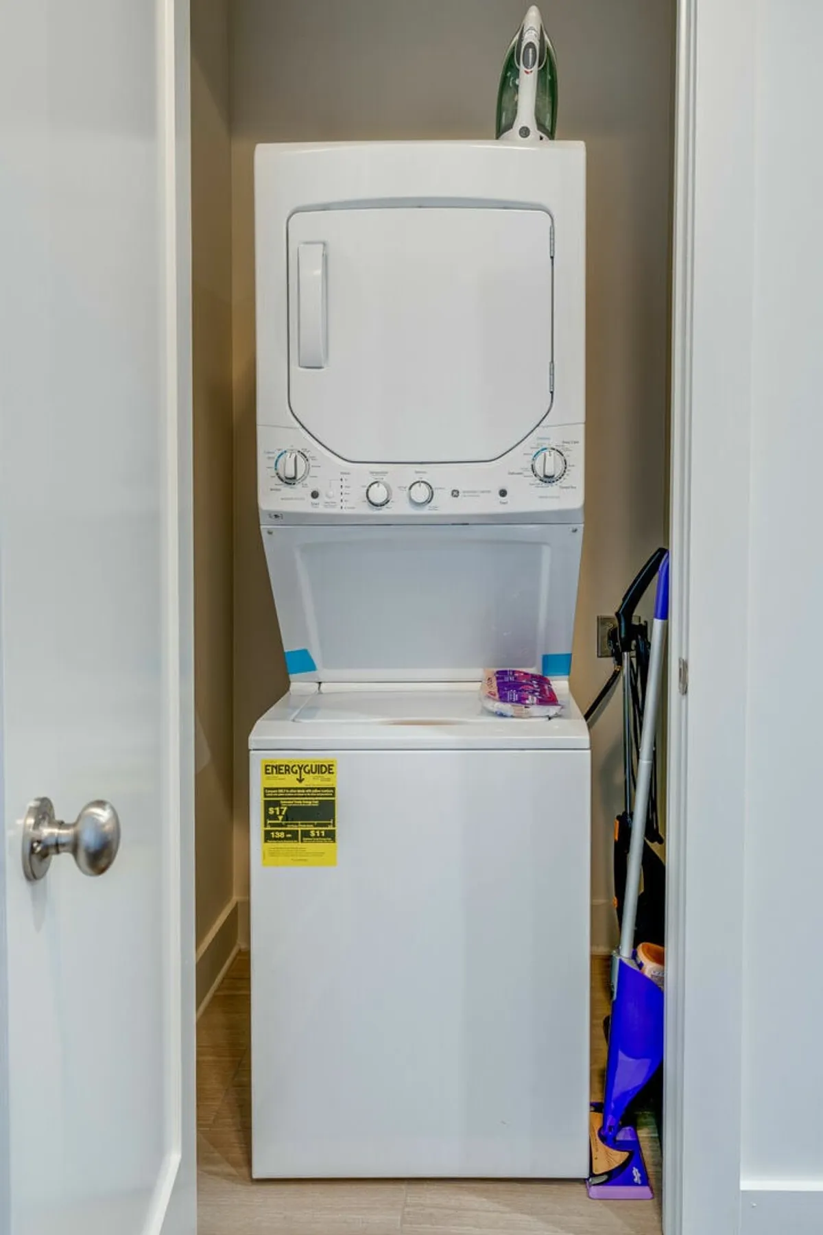 A stacked washer and dryer set is neatly integrated into a compact nook, with an iron placed on top and cleaning supplies in the corner. The space is tidy and efficient, emphasizing practical in-unit amenities.